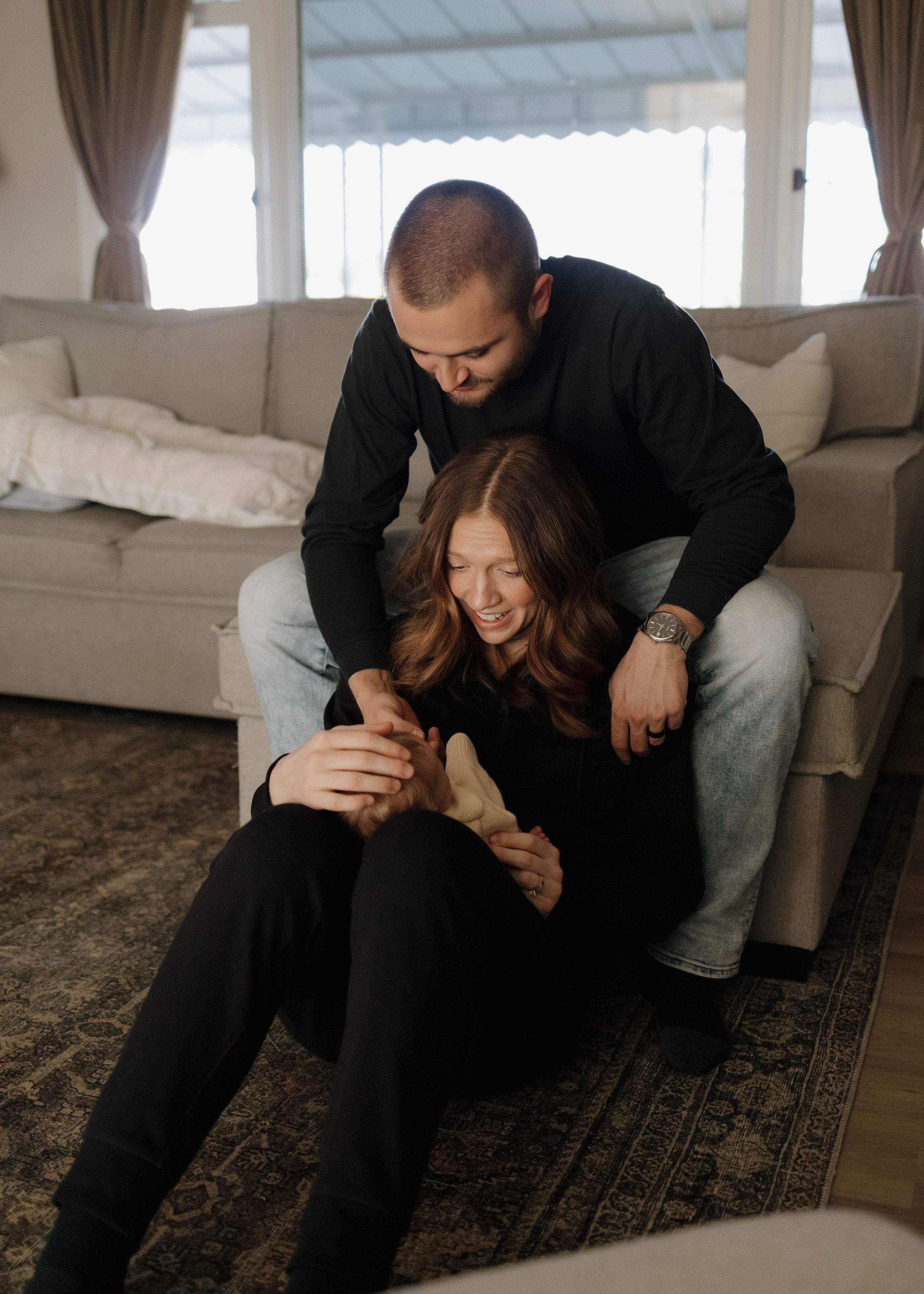Couple sharing a playful intimate moment while sitting together on beige couch in sunlit living room with patterned rug.