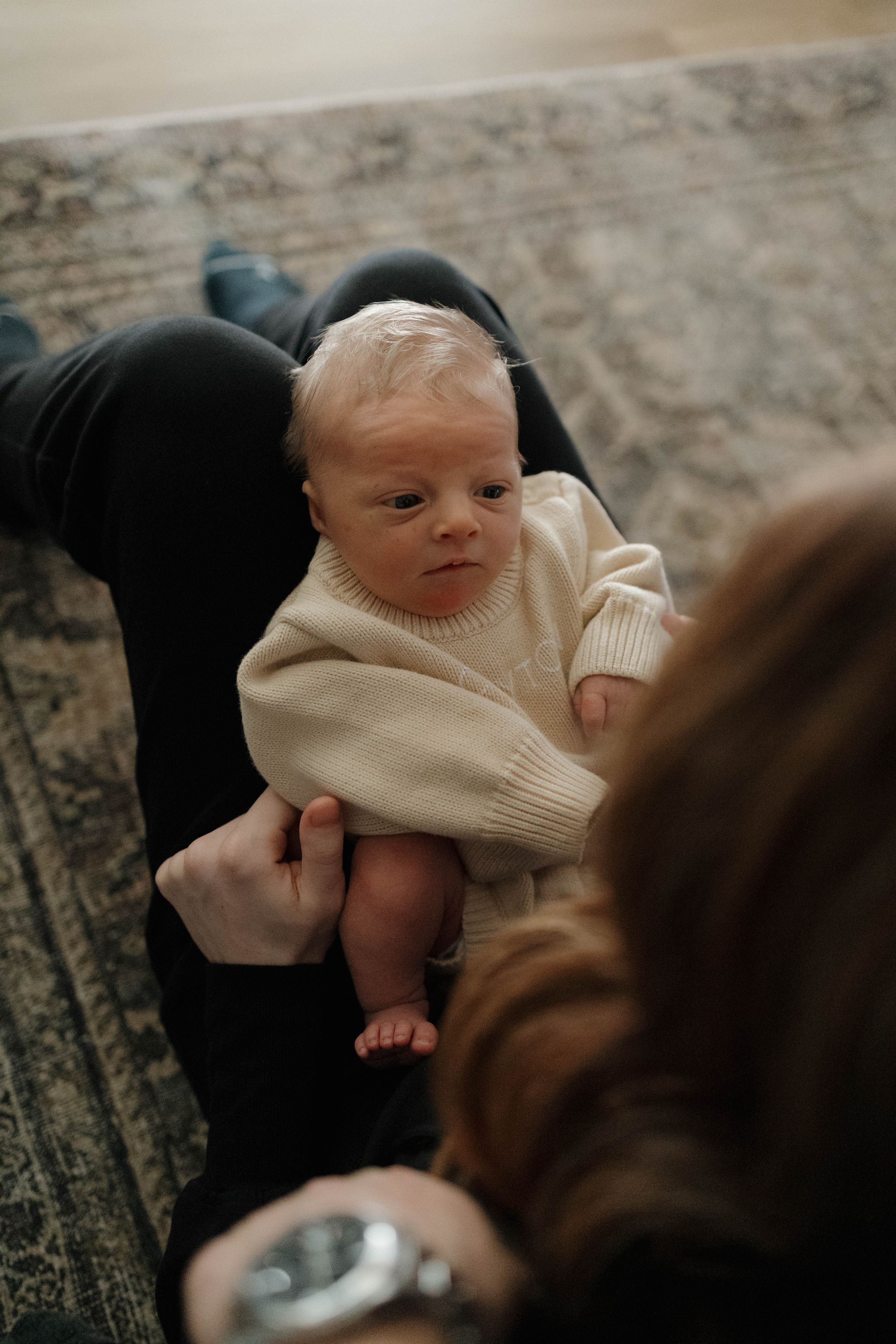 A baby wearing a cozy cream sweater rests on a patterned carpet while being held by adults.