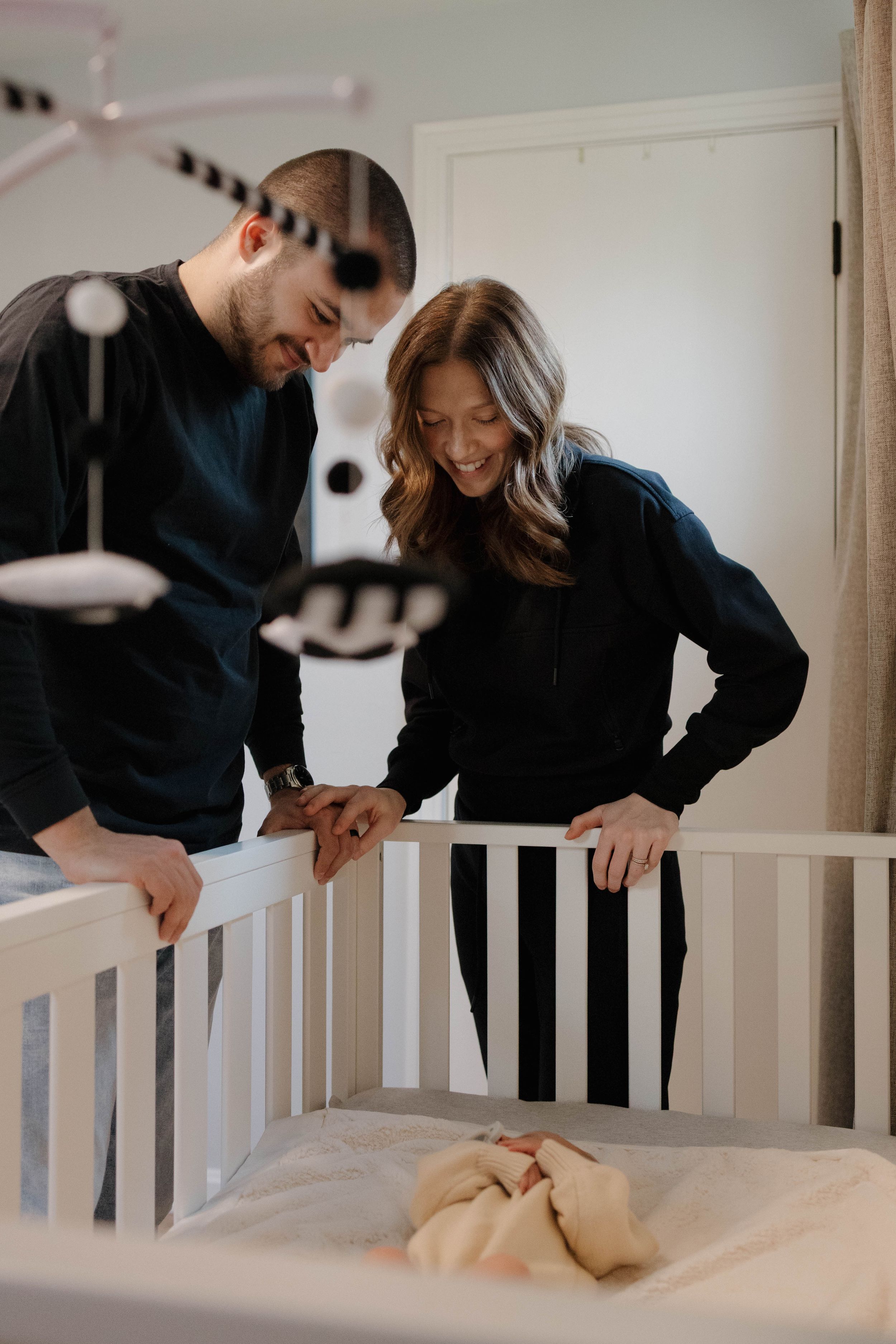 A couple looks down lovingly at their baby's white crib in a cozy, dimly lit nursery.