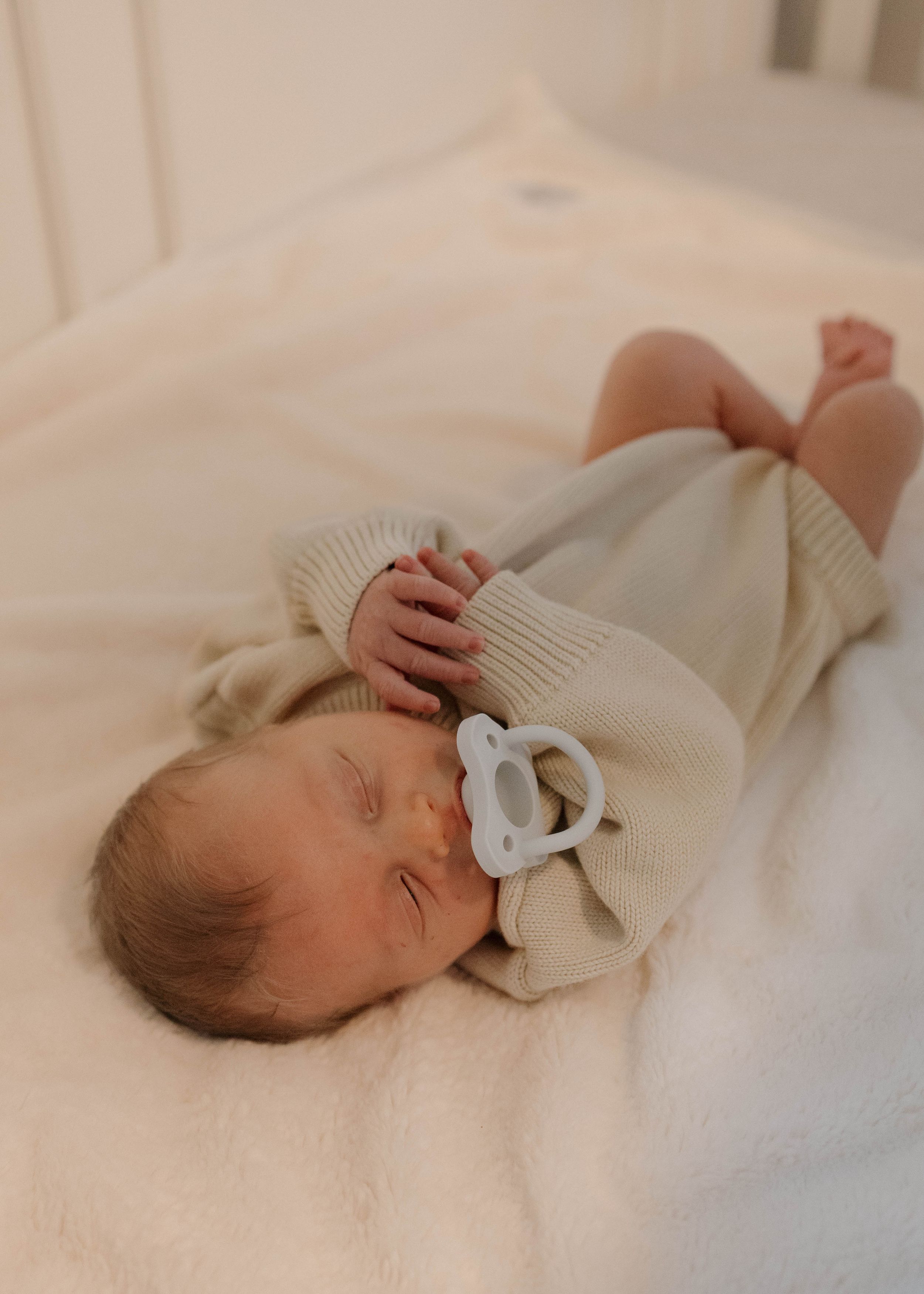 Sleeping infant wearing cream knit sweater rests peacefully on white bedding.
