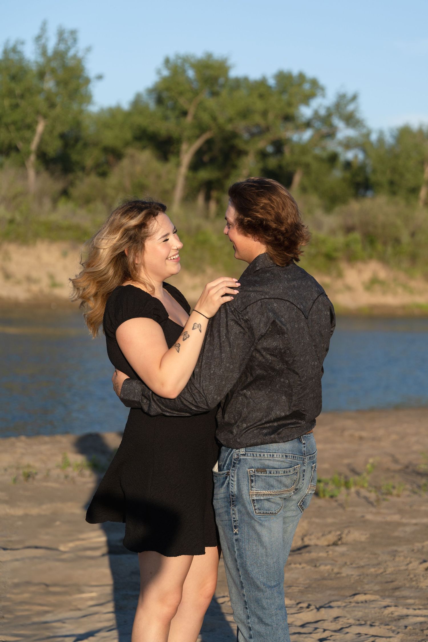 A romantic couple shares a tender moment while dancing together on a beach shore at sunset by a lake.