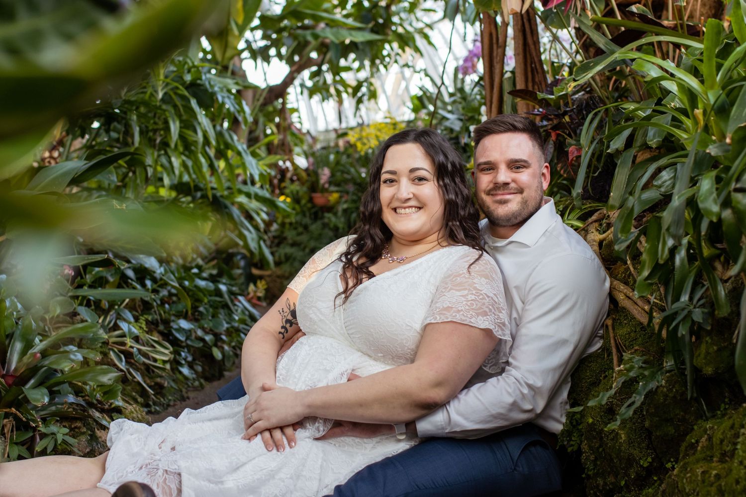 A couple poses together outdoors in casual attire surrounded by lush greenery and tropical plants.