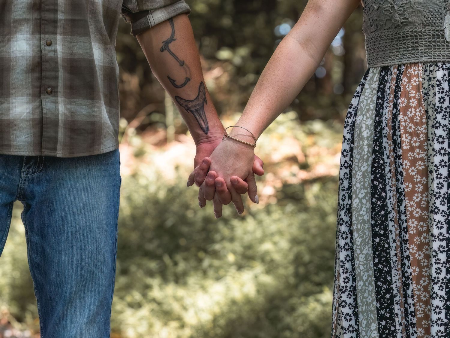 Two people hold hands intimately in a garden setting, one wearing jeans and one wearing a patterned skirt.