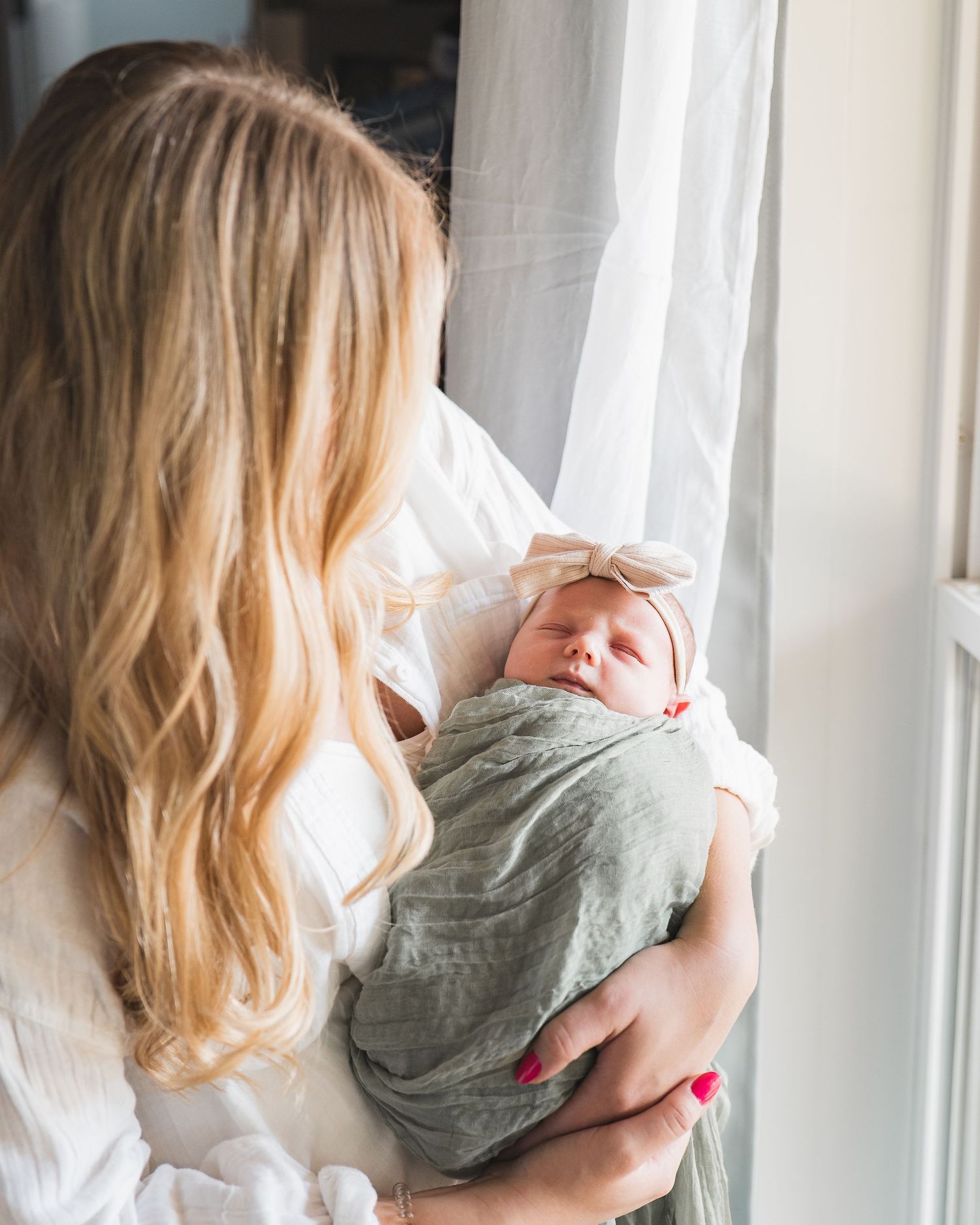A woman in white holds a sleeping newborn wrapped in sage green swaddle next to a bright window.