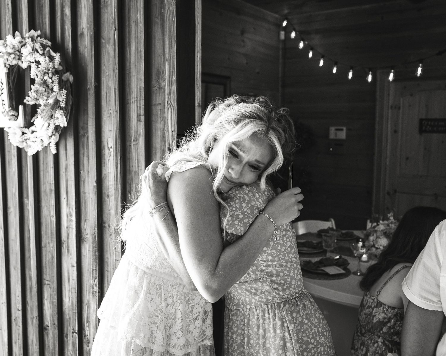 Two people in light colored dresses share an emotional embrace at an indoor venue with string lights and decor.