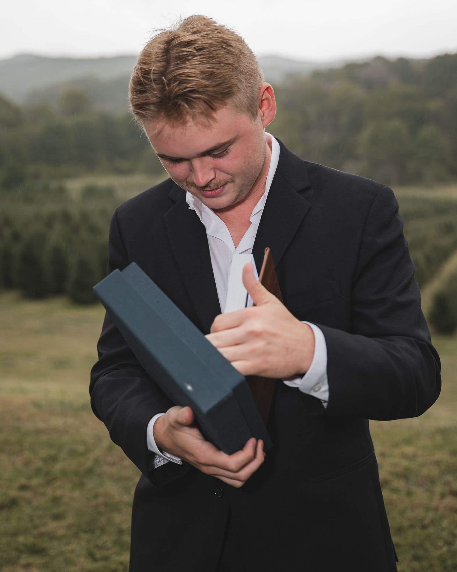 Person in formal attire smiling while holding a dark blue package outdoors.