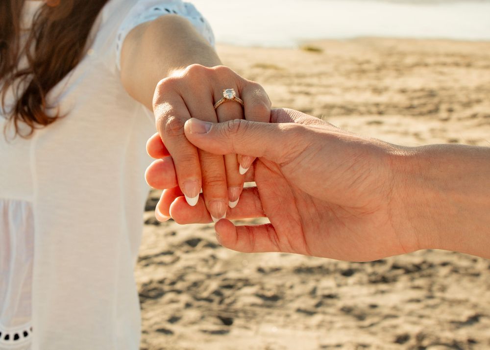 Close-up of hands displaying an engagement ring on a sunny beach.