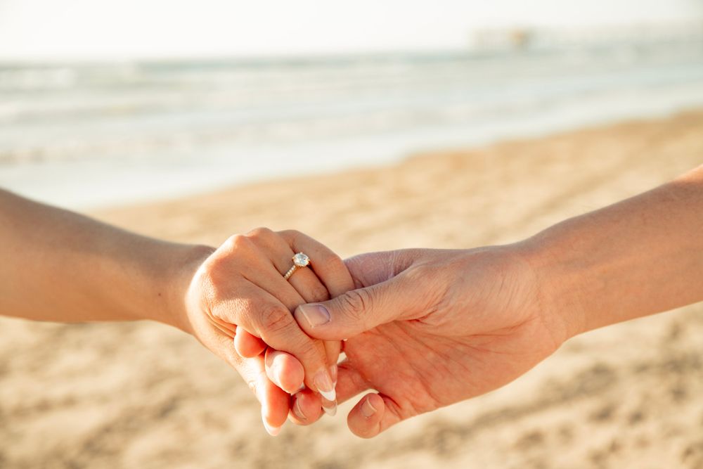 Close-up shot of two hands clasped together on a sandy beach at sunset.