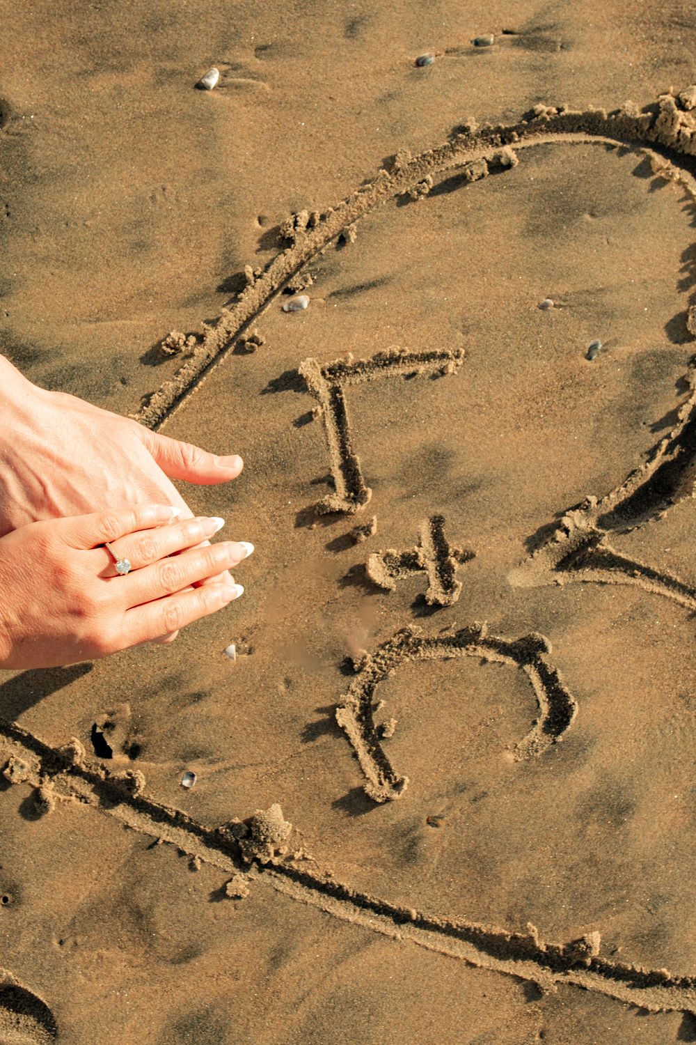 A heart drawn in wet sand on the beach with hands reaching to complete it.