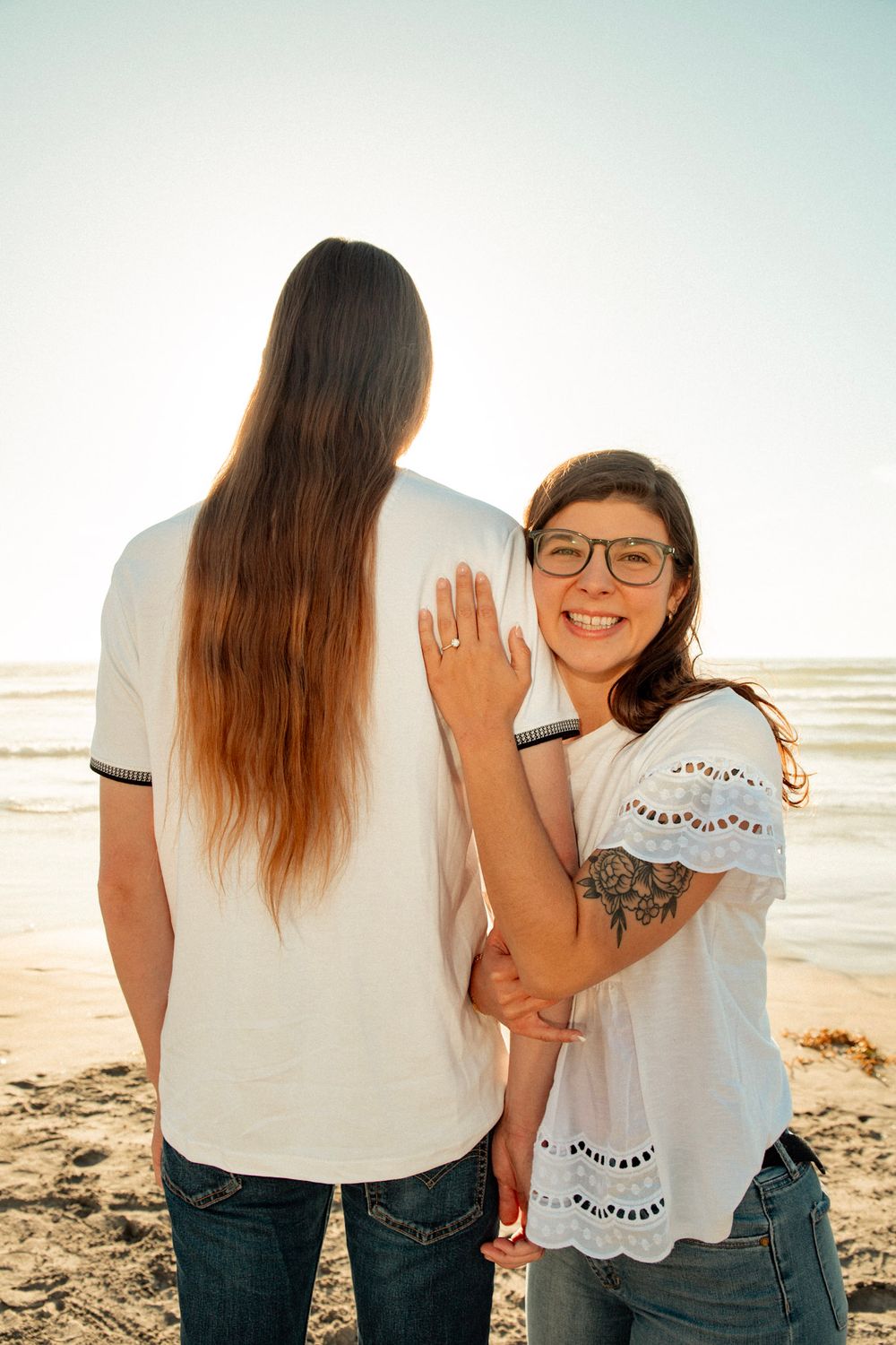 Couple in matching white shirts on beach showing off engagement ring.