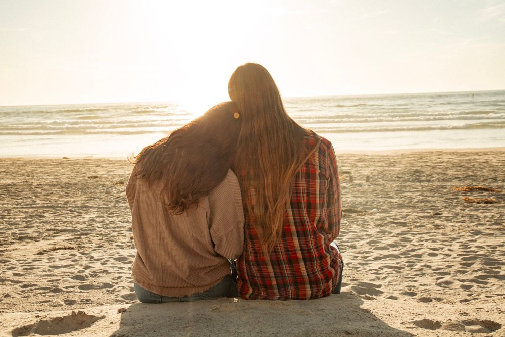 Back view of couple in casual wear sitting close together on beach at sunset.