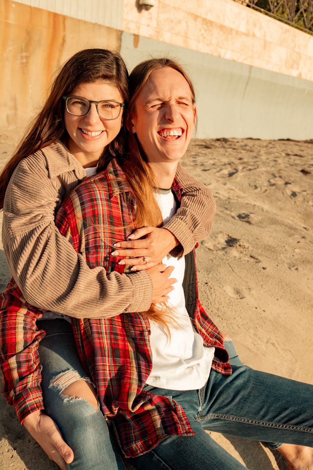 Couple sharing joyful laughter while embracing on beach during golden hour.