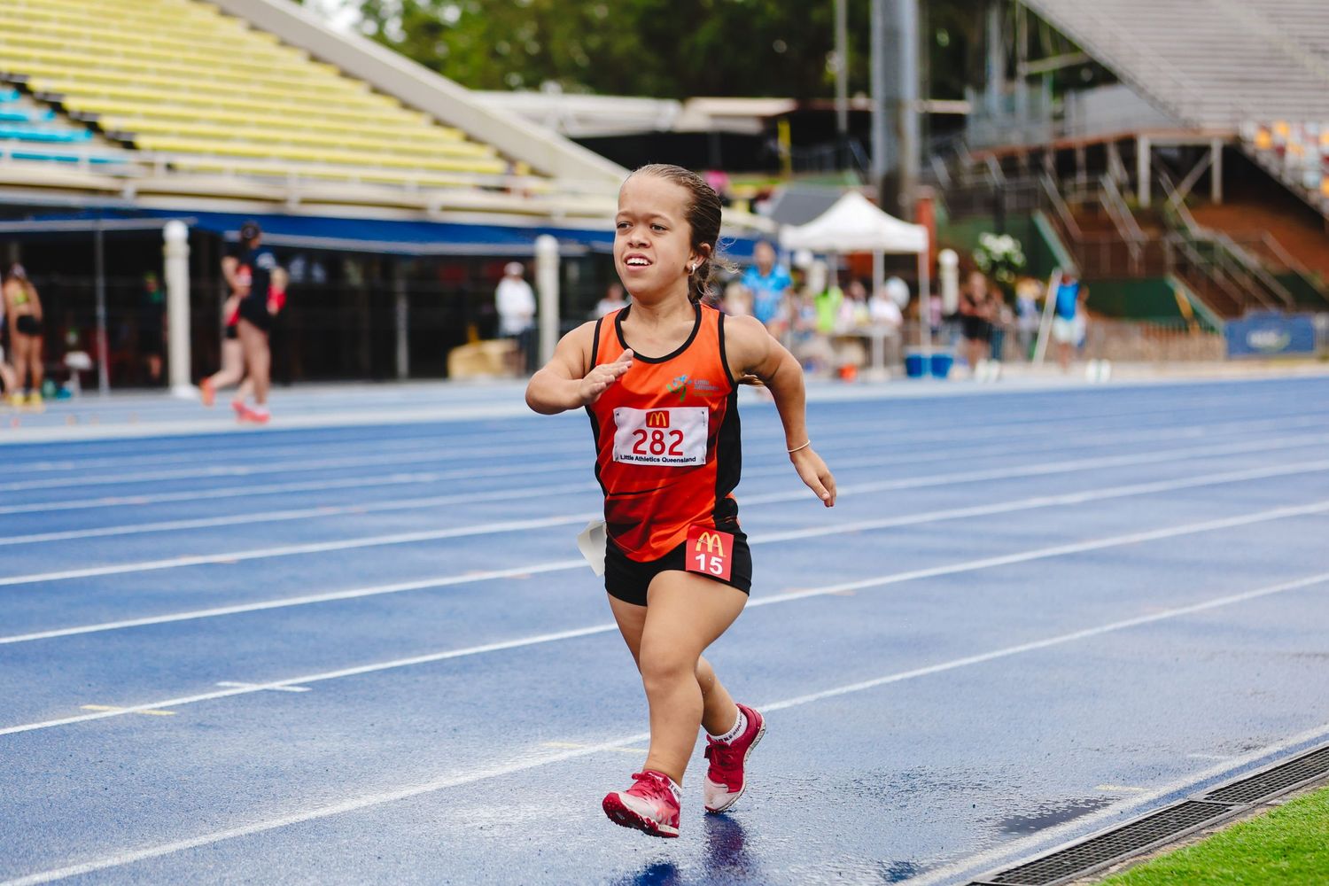 A runner in red jersey and black shorts competes on a blue track at an outdoor athletics stadium.