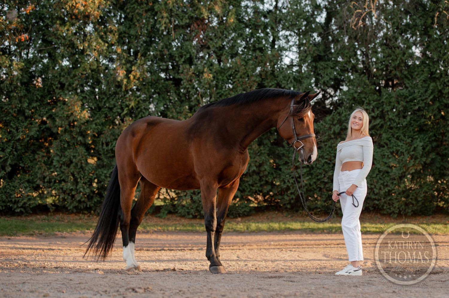 A bay horse stands alert next to someone in all-white attire against a backdrop of autumn trees.