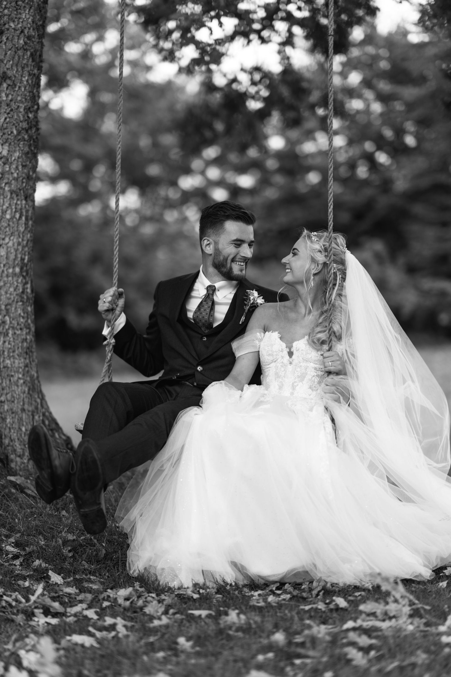 A bride and groom share a joyful moment while sitting together on a rustic tree swing in a romantic forest setting.