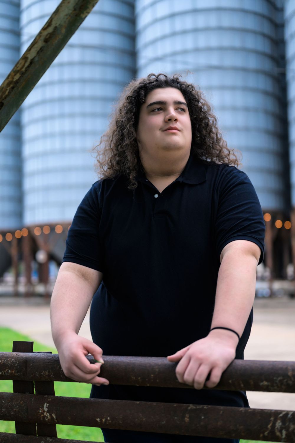 Person with long curly dark hair wearing a black polo shirt leans against railing with industrial silos in background.
