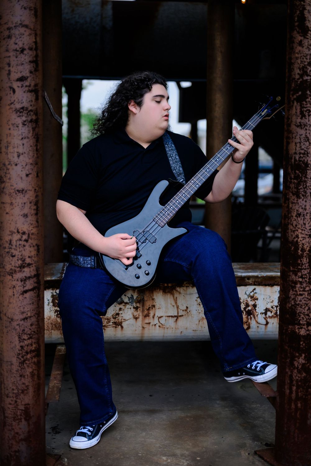 Musician playing electric bass guitar while sitting in industrial setting wearing black shirt and blue jeans.