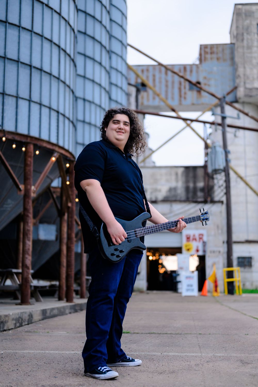 A musician holding a guitar poses in front of an industrial building with curved glass walls and metal beams.