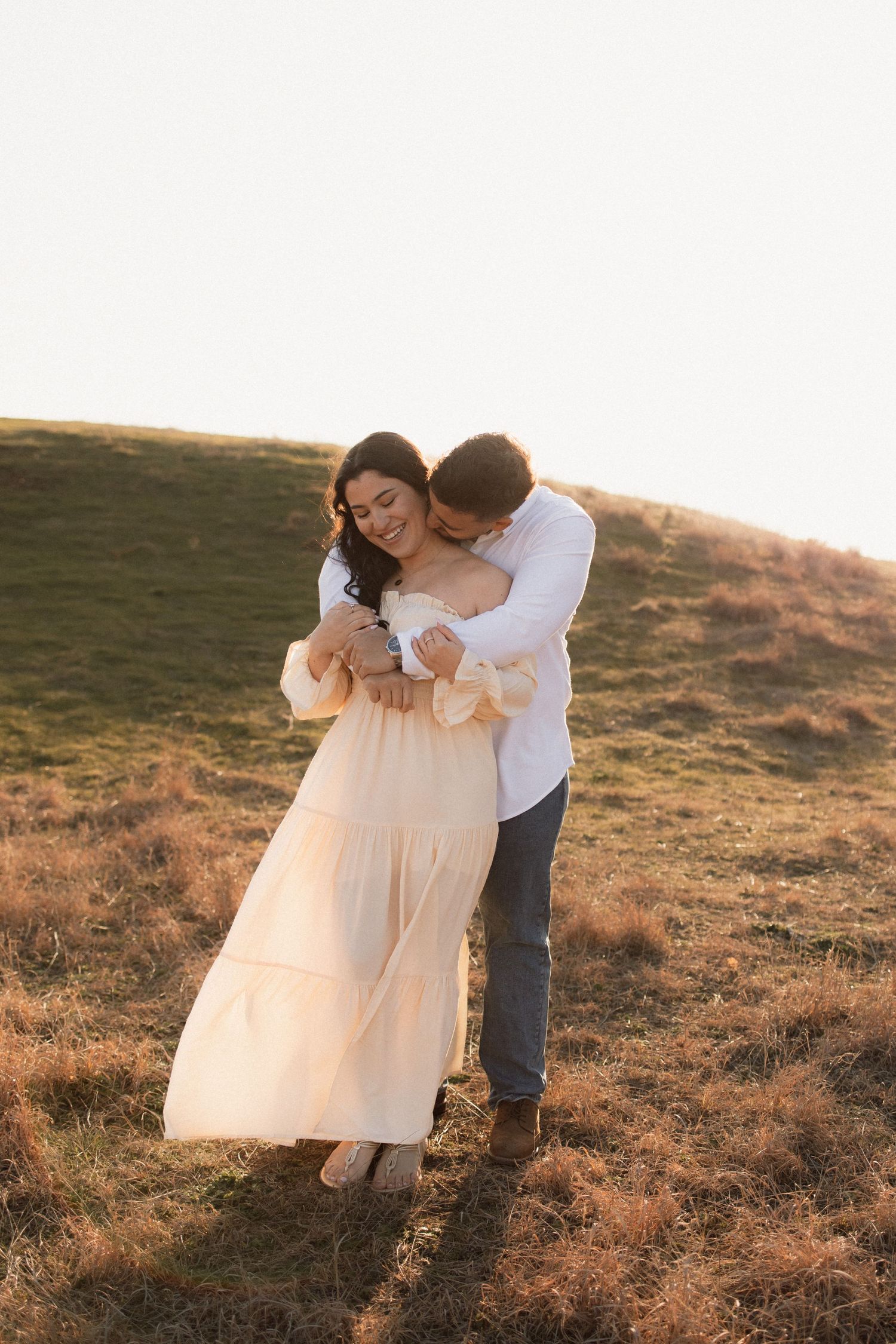 A romantic couple embraces on a hillside at sunset, with the woman wearing a flowing cream-colored maxi dress.