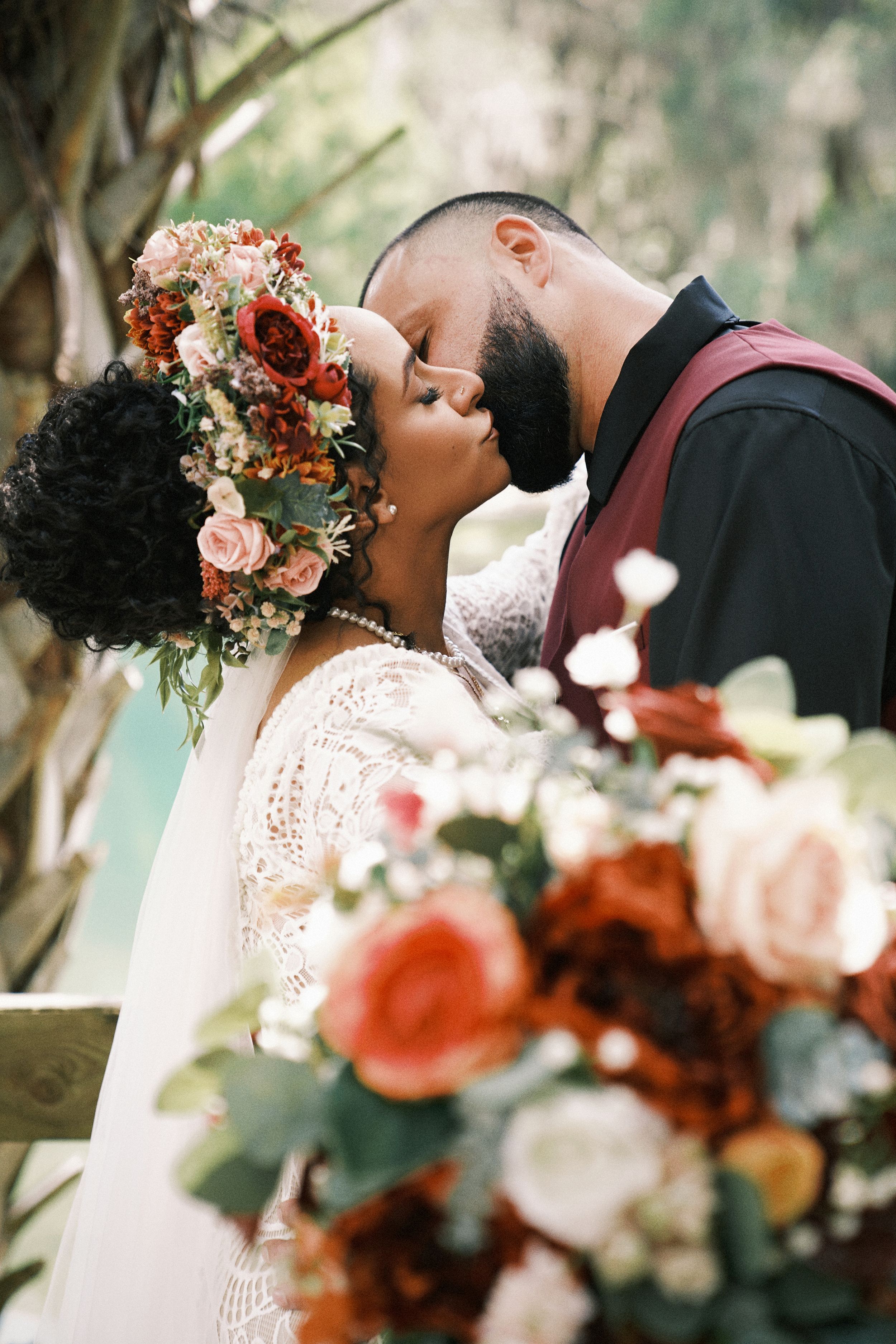 Bride and groom sharing a kiss surrounded by lush floral arrangements with red and pink blooms at their wedding ceremony.