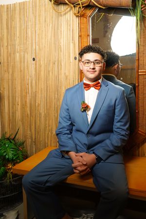 Person in a blue suit and red bow tie sitting on a bench against bamboo wall decor.