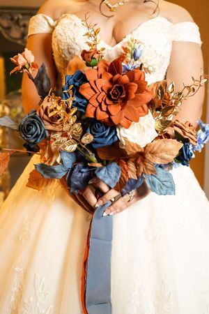 Rustic autumn bridal bouquet featuring navy blue, rust orange and white flowers with gold accents against a white wedding gown.