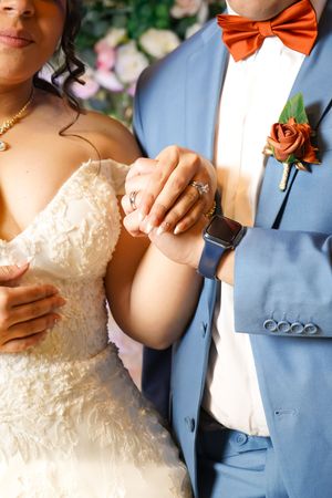 Close-up wedding photo showing hands with rings, light blue suit with orange bowtie, and white lace dress details.
