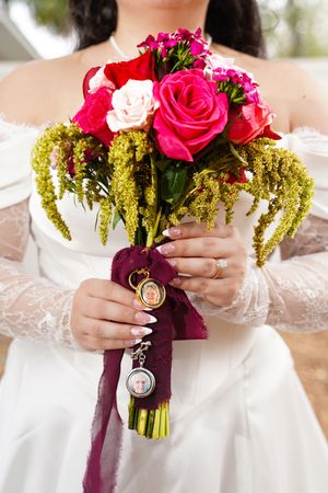 Wedding bouquet with vibrant pink and red roses wrapped in a dark purple ribbon with vintage locket attachments.