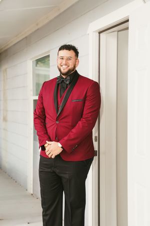 A person in a stylish burgundy suit jacket and black dress pants poses confidently against a white wall.