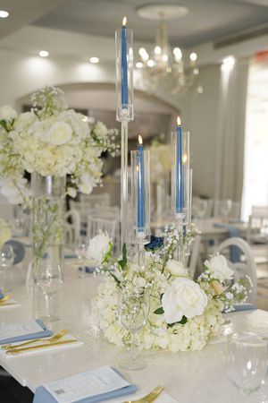 Wedding reception table settings with white floral centerpieces, blue candles, and elegant place settings in a banquet hall.