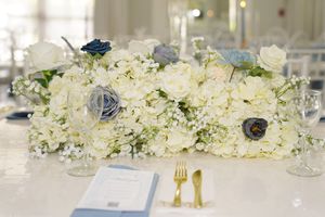 An elegant white hydrangea and blue flower wedding centerpiece arrangement with gold utensils and menu card on table.
