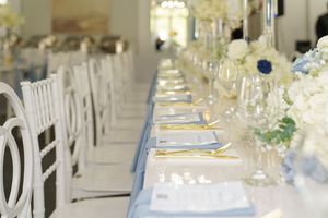 Elegant white wedding reception tables decorated with white floral centerpieces and blue accents in a bright venue.