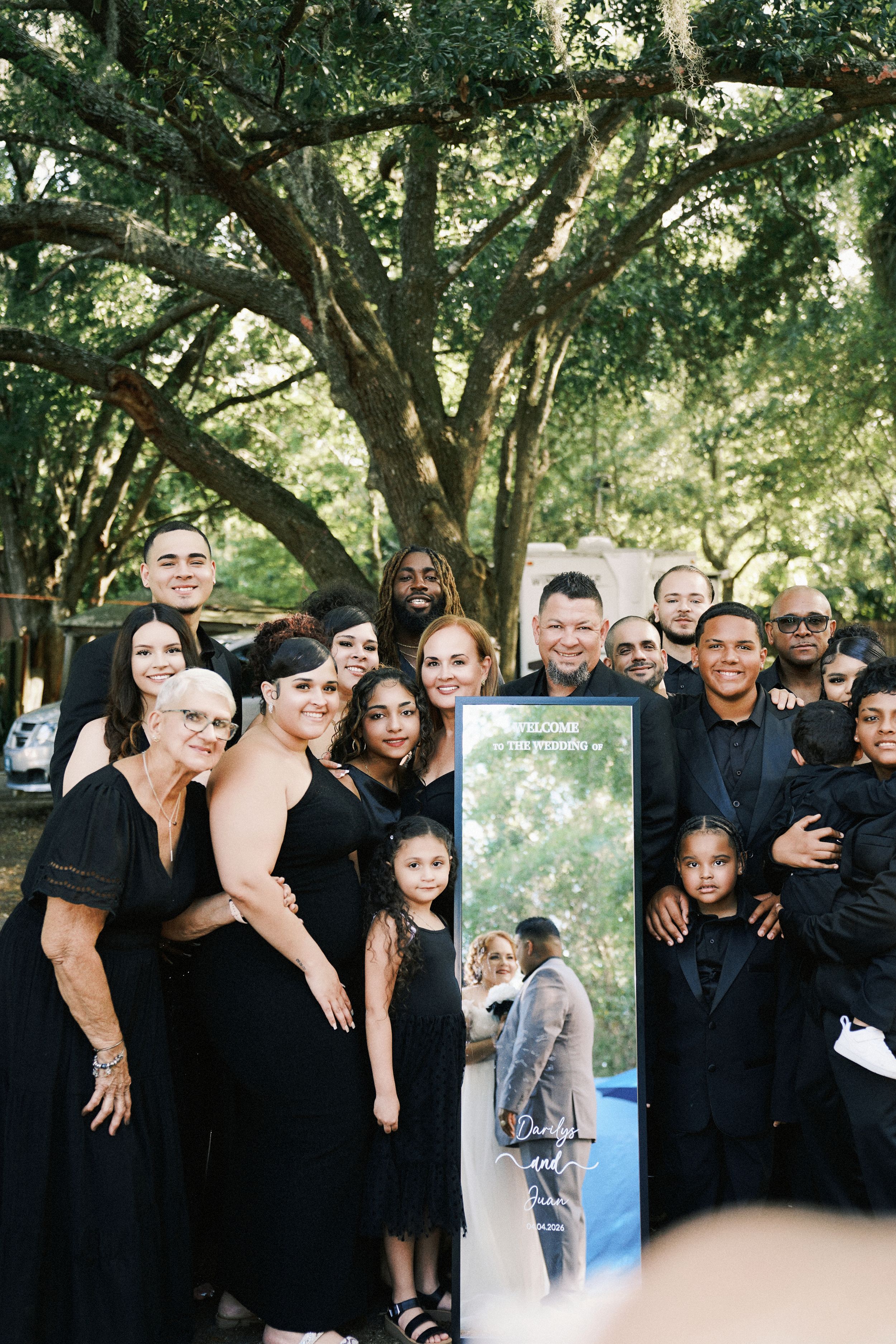 Large wedding party group photo in black attire posing outdoors under oak trees with a decorative mirror sign.