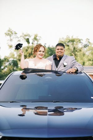 Bride and groom smiling through sunroof of luxury car, bride holding bouquet, both dressed in wedding attire outdoors.