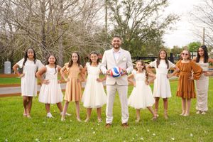 Group photo of wedding party members in coordinating white and brown dresses arranged on grassy outdoor setting.