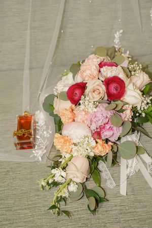 A bridal bouquet with pink and peach roses, red ranunculus, white hydrangeas and eucalyptus leaves next to a perfume bottle.