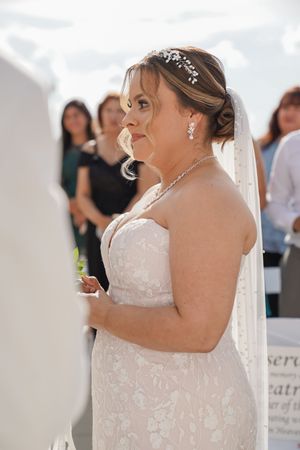 A bride wearing a strapless white lace wedding dress with delicate jewelry and a veil smiles during the ceremony.