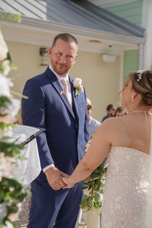 A wedding ceremony outdoors with two people exchanging vows beneath a white-columned entrance with floral decorations.