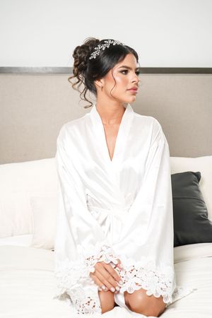 Woman in white silk and lace bridal robe with crystal headpiece sits elegantly on couch looking to side.