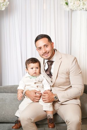 A parent and small child wearing matching beige suits sit together on a gray couch with white floral decor in background.