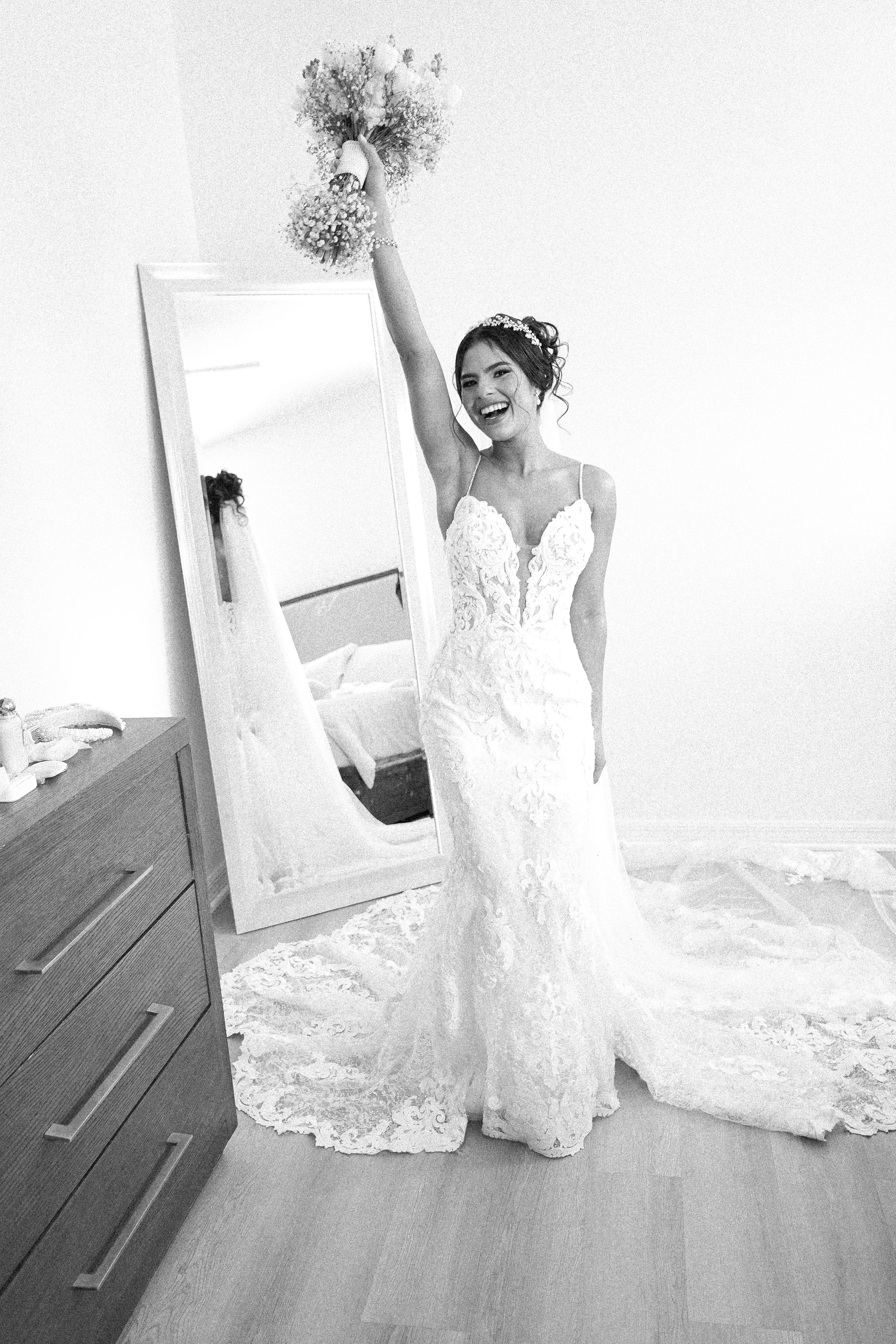 A bride wearing a white lace wedding gown with sweetheart neckline poses joyfully near a full-length mirror.