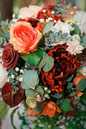 Bridal bouquet with orange and burgundy roses, eucalyptus, baby's breath, and a gold locket charm with a photo memorial.