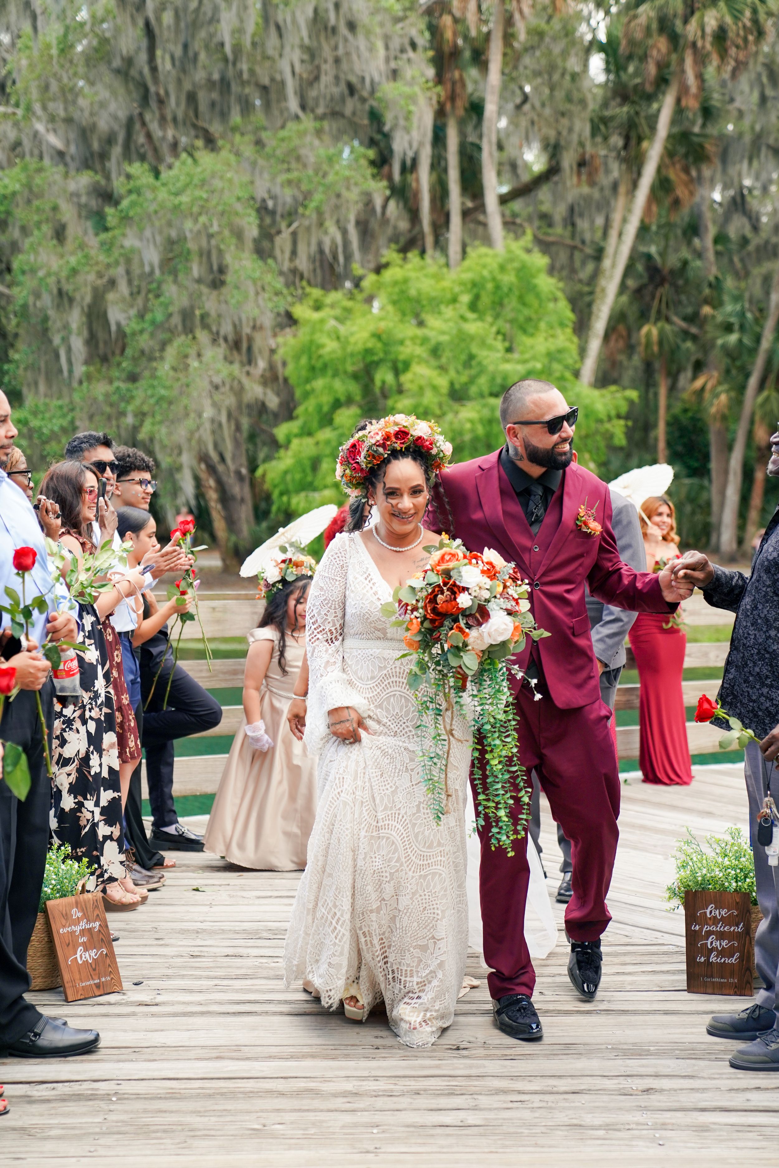 Bride in lace gown with floral crown walks the aisle with groom in burgundy suit on outdoor wooden walkway.