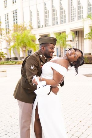 A couple in formal attire, with one in military dress uniform and one in a white gown, share a romantic dip pose outdoors.