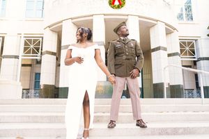 A couple poses on steps of a building, with one in military dress uniform and one in an elegant white dress with a slit.