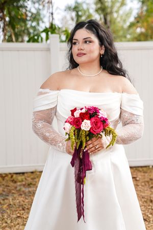 Elegant off-shoulder white wedding dress with lace sleeves and vibrant red and pink rose bouquet in outdoor setting.