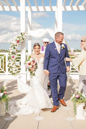 Elegant wedding ceremony takes place under white pergola decorated with pink flowers on sunny day with cloudy sky.