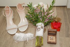 Elegant white bridal heels displayed alongside decorative greenery, rings, and red floral accents on a wooden floor.