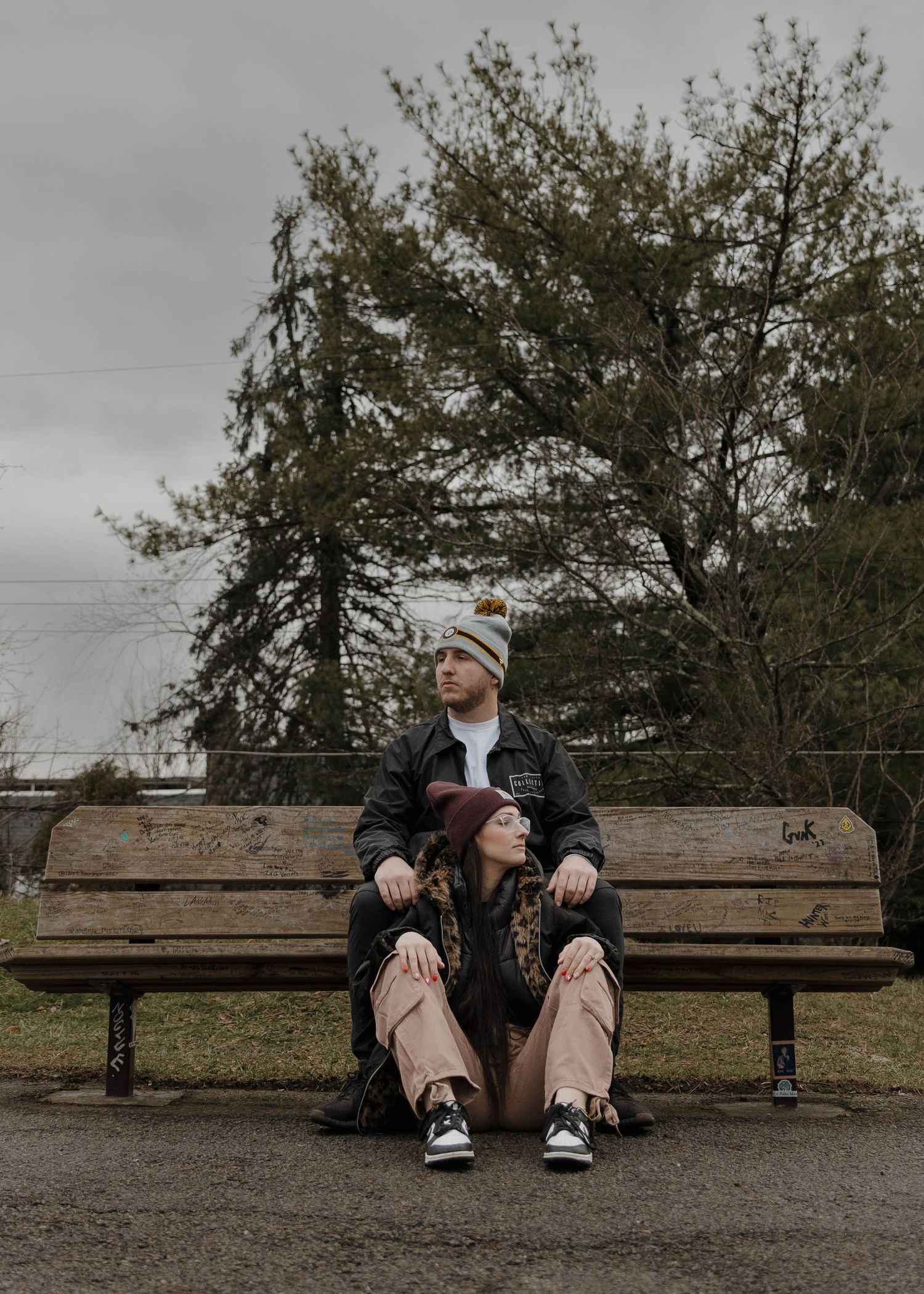 A person sits alone on a wooden bench against a backdrop of tall trees on a cloudy day.