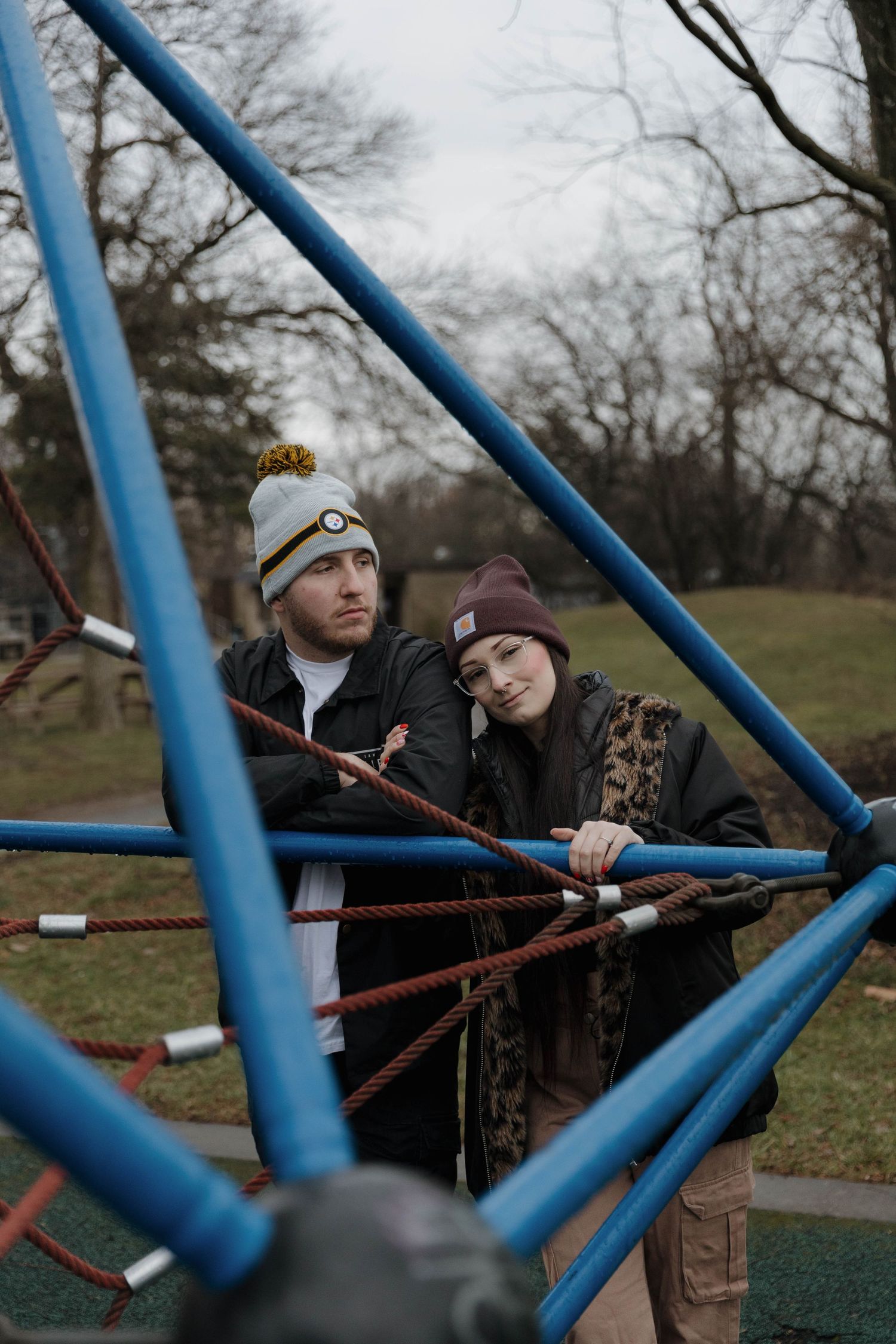 A couple leans against an outdoor climbing dome structure with blue metal bars on a cloudy winter day.